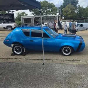 Blue custom AMC Gremlin with black racing stripe and hood scoop at an outdoor car show.
