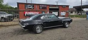 Black classic Camaro SS parked in front of the orange JR Muffler & Mechanic Shop building in Palestine, Texas, with signs for full-service mechanic work, shocks and struts.