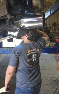 Mechanic in a welding helmet and a JR Muffler and Mechanic Shop shirt listing brakes, suspension and A/C, welding a polished exhaust tip under a truck on a lift.