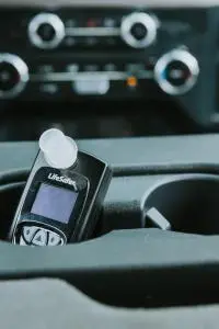 A black LifeSafer ignition interlock device with a white mouthpiece rests in a vehicle's center console cup holder, with the dashboard blurred in the background.